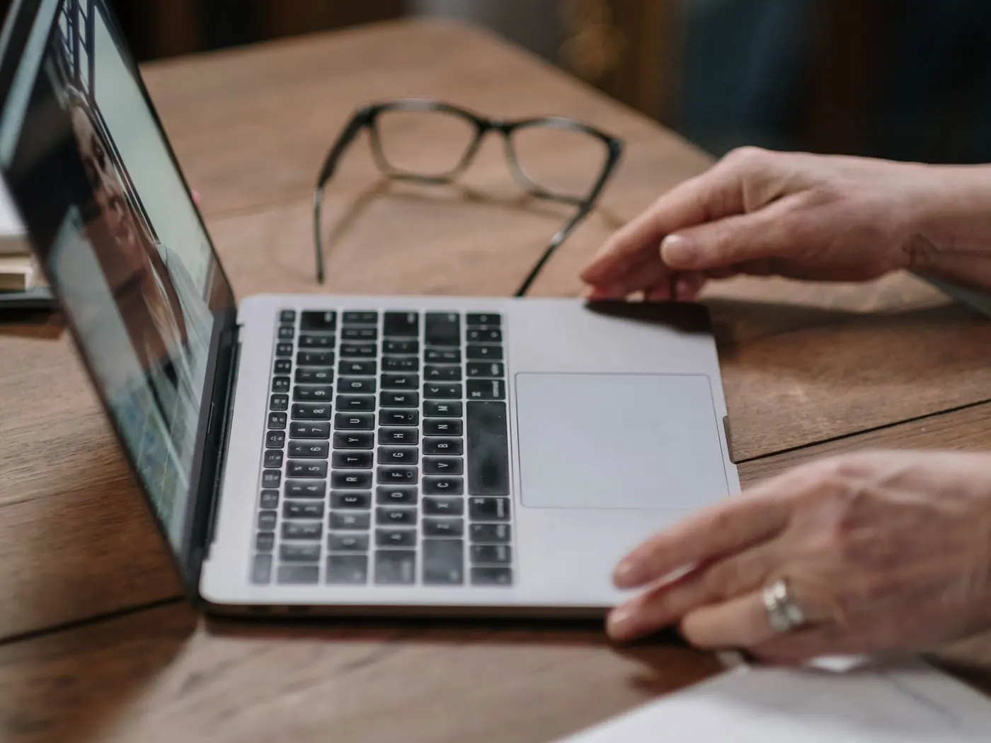 Manos sobre el teclado de un portátil durante una videollamada, con unas gafas y unos papeles al lado