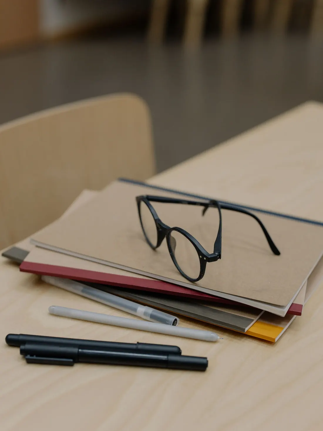 Carpetas apiladas con unas gafas encima y varios bolígrafos sobre una mesa de trabajo